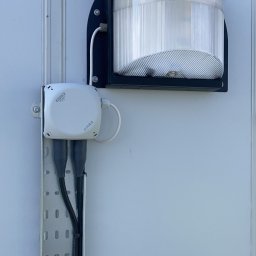 Andrzej Korda - Zewnętrzna lampa z czarną obudową i przezroczystym kloszem, zamontowana na białej ścianie. Widoczna puszka elektryczna i kable w metalowej listwie. Oświetlenie elewacyjne.