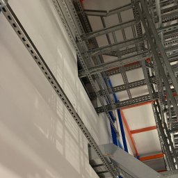 Andrzej Korda - Metalowe korytka kablowe zamontowane pod sufitem, widok z dołu, z perspektywą na konstrukcję nośną. Szare elementy na tle białej ściany i sufitu.