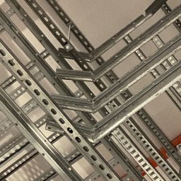 Andrzej Korda - Metalowe korytka kablowe, perforowane, zamontowane pod sufitem, tworzące skomplikowaną sieć. Widoczne elementy mocujące i fragmenty pomarańczowych rur.