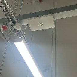 Andrzej Korda - Zamontowana lampa LED na suficie podwieszanym, widoczne elementy instalacji elektrycznej i rury. Oświetlenie biurowe, sufit kasetonowy.