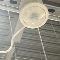 Andrzej Korda - Zamontowany, okrągły czujnik ruchu w białej obudowie, przymocowany do metalowej szyny pod sufitem. Widoczny przewód. Ujęcie z dołu.