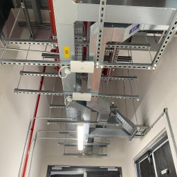 Andrzej Korda - Sufit z widocznymi, zamontowanymi korytami kablowymi, przewodami i elementami oświetlenia. Widoczne kable w kolorze czerwonym. Ujęcie od dołu.