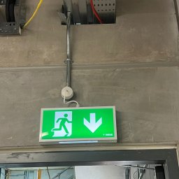 Andrzej Korda - Montaż oświetlenia awaryjnego: zielony znak ewakuacyjny z podłączeniem elektrycznym na szarej ścianie, widoczne kable i metalowe korytka instalacyjne.