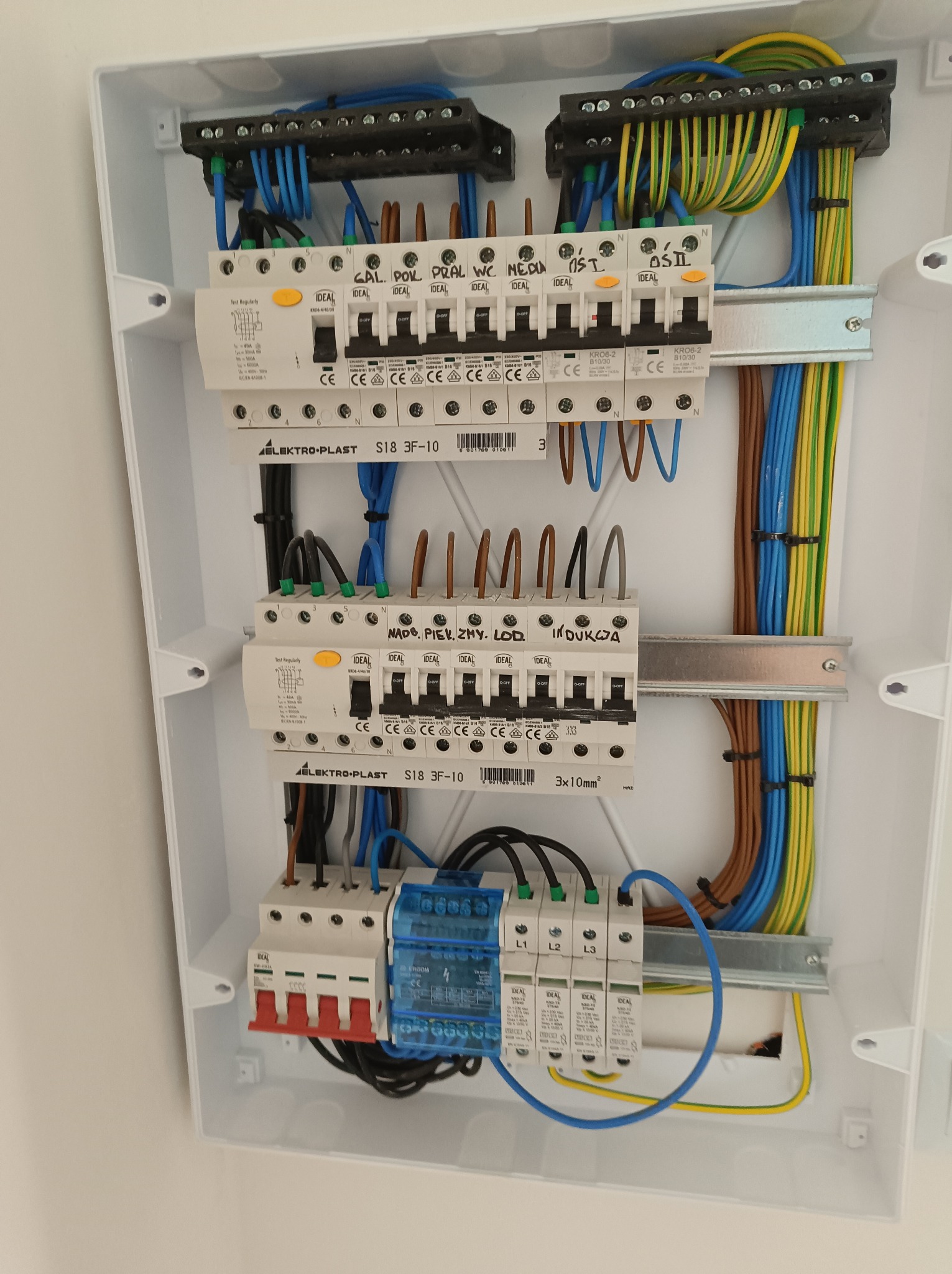 Rozdzielnica elektryczna w białej obudowie, z widocznymi bezpiecznikami, przewodami i listwami zaciskowymi. Uporządkowane okablowanie wewnątrz skrzynki.