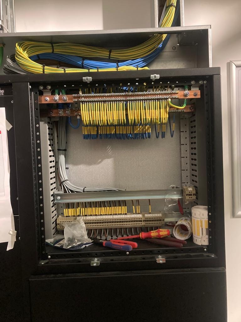 Rozdzielnica elektryczna z podłączonymi przewodami w kolorach żółto-zielonym, niebieskim i szarym. Widoczne narzędzia i akcesoria instalacyjne.