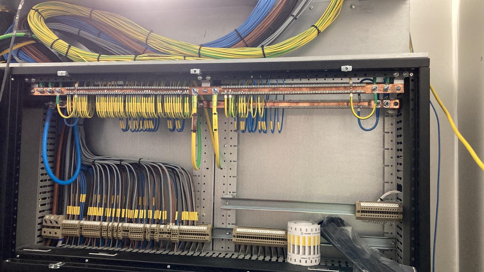 Rozdzielnia elektryczna z precyzyjnie ułożonymi kablami w różnych kolorach, podłączonymi do listew zaciskowych i szyn wyrównawczych. Widoczne oznaczenia na przewodach.