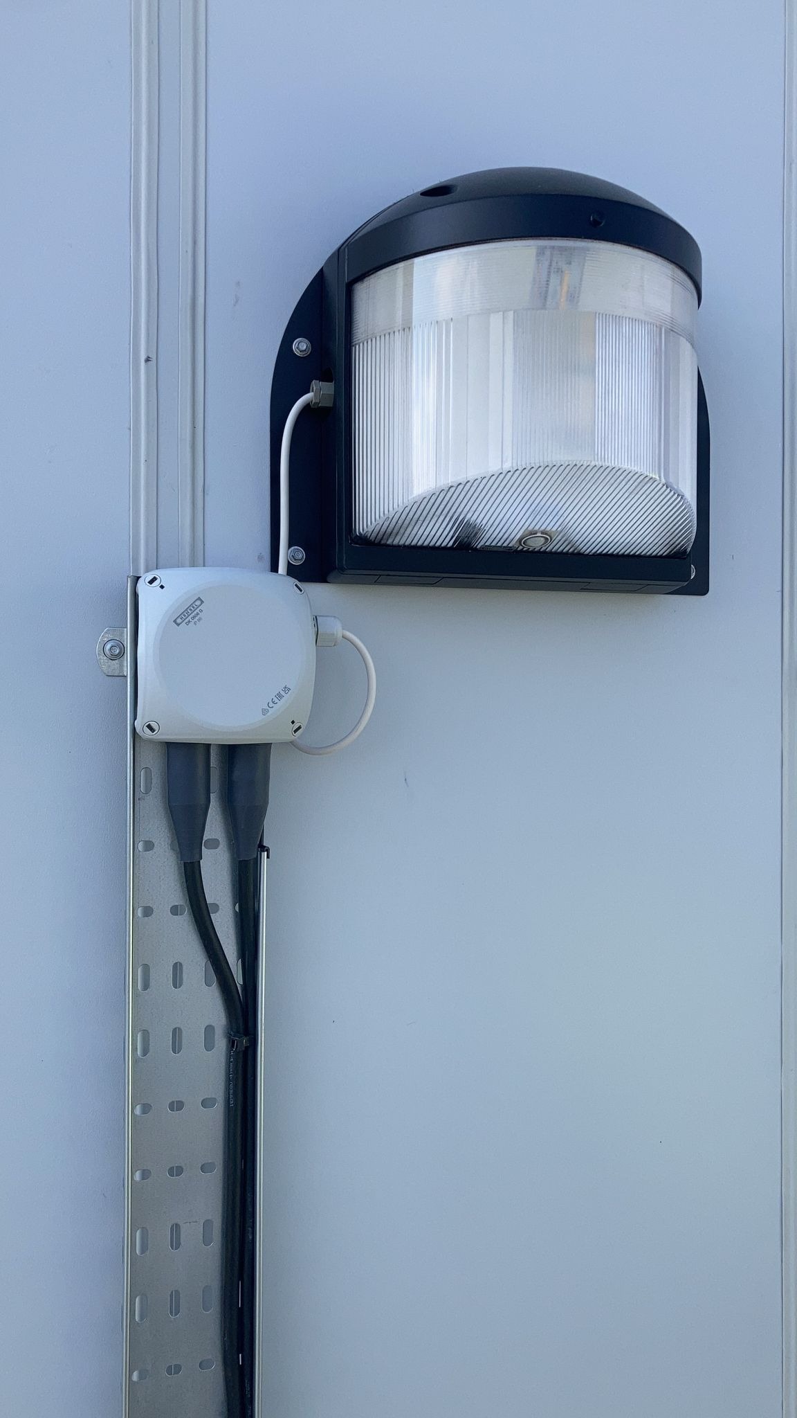Zewnętrzna lampa z czarną obudową i przezroczystym kloszem, zamontowana na białej ścianie. Widoczna puszka elektryczna i kable w metalowej listwie. Oświetlenie elewacyjne.