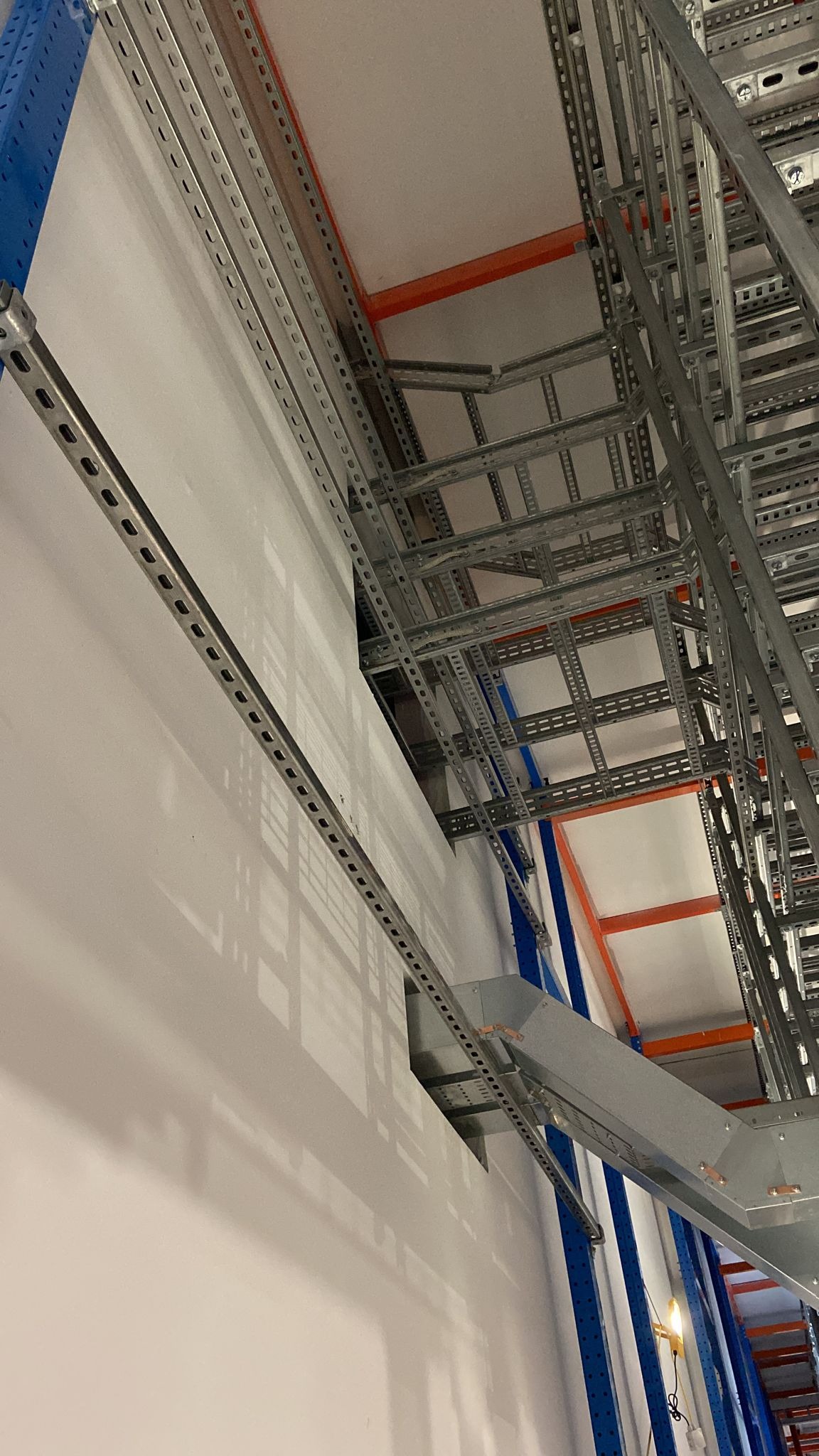 Metalowe korytka kablowe zamontowane pod sufitem, widok z dołu, z perspektywą na konstrukcję nośną. Szare elementy na tle białej ściany i sufitu.