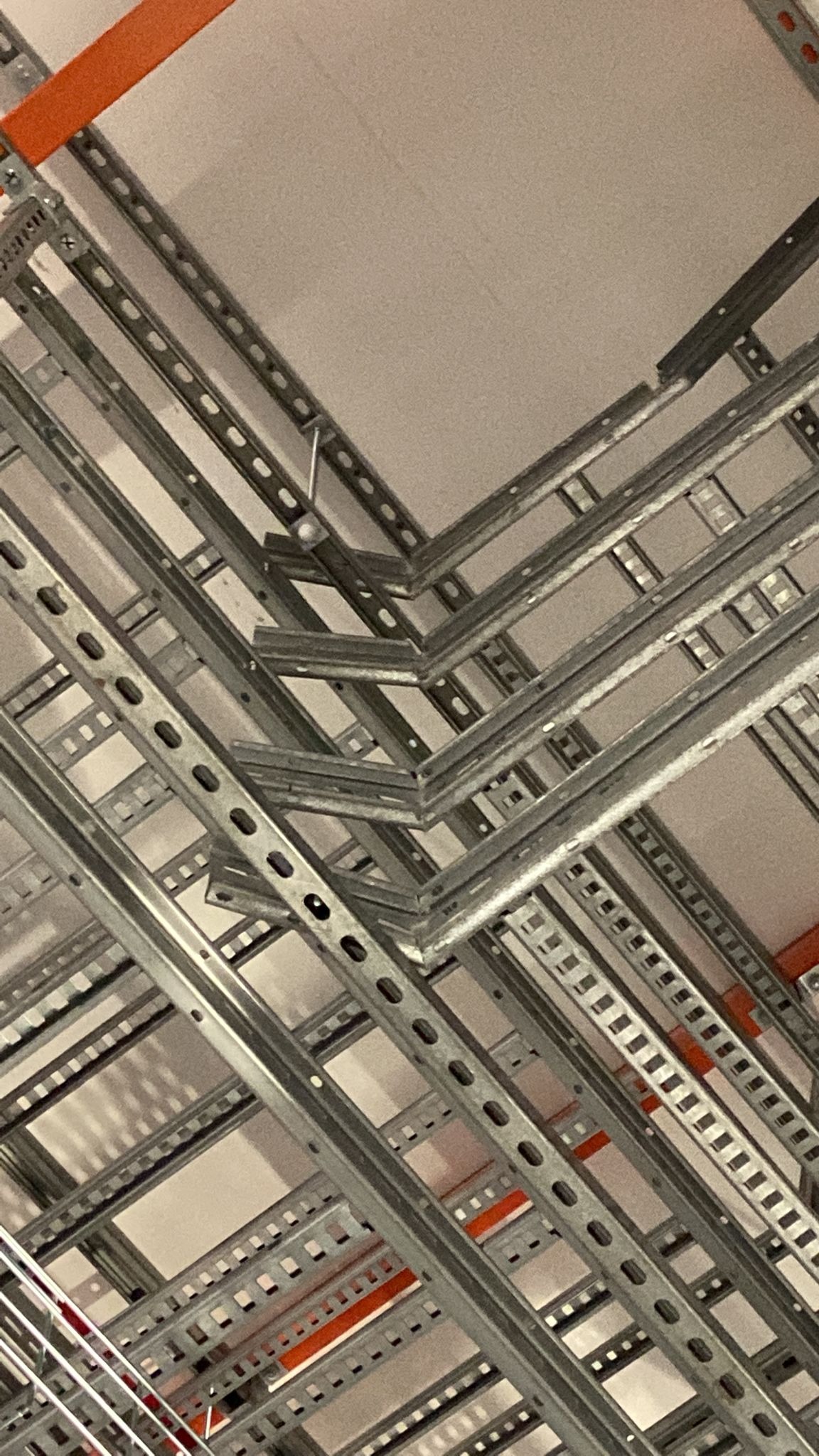 Metalowe korytka kablowe, perforowane, zamontowane pod sufitem, tworzące skomplikowaną sieć. Widoczne elementy mocujące i fragmenty pomarańczowych rur.