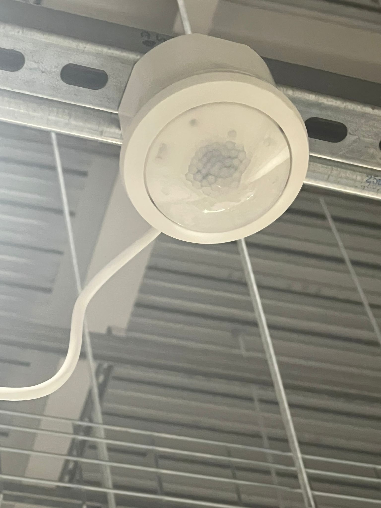 Zamontowany, okrągły czujnik ruchu w białej obudowie, przymocowany do metalowej szyny pod sufitem. Widoczny przewód. Ujęcie z dołu.