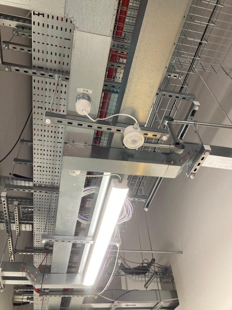 Sufit z zamontowanymi metalowymi korytkami kablowymi, oświetleniem LED i czujnikiem ruchu PIR. Widoczne przewody w kolorowych izolacjach.