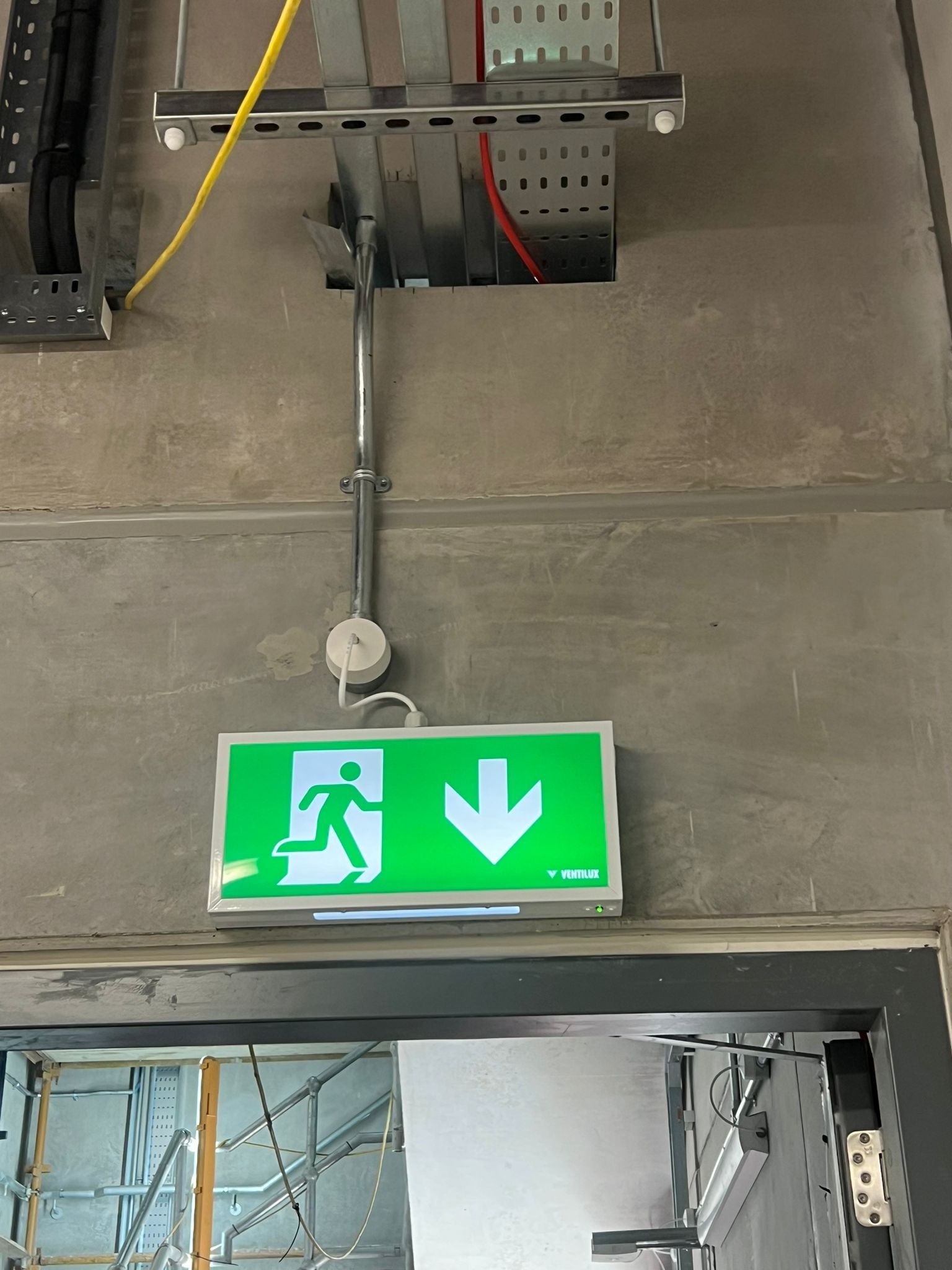 Montaż oświetlenia awaryjnego: zielony znak ewakuacyjny z podłączeniem elektrycznym na szarej ścianie, widoczne kable i metalowe korytka instalacyjne.