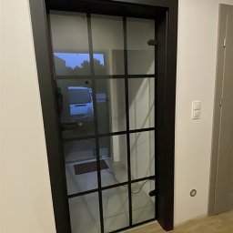 KO-LEŚ-BUD - Czarne, przeszklone drzwi wewnętrzne z kratownicą, widoczne narzędzia i chemia budowlana na podłodze. W tle widoczny samochód dostawczy.