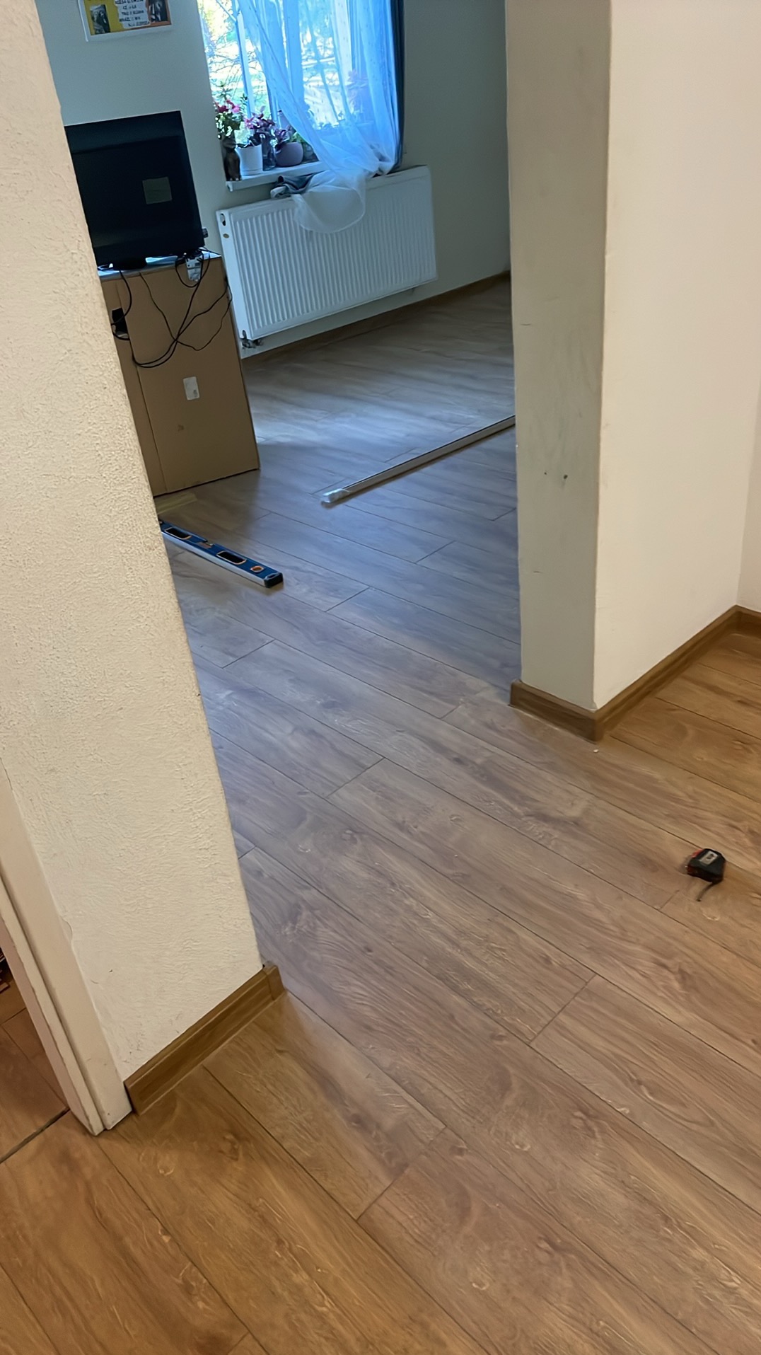 Ukończona podłoga z paneli w odcieniach brązu, widoczna przy ścianie listwa przypodłogowa, poziomica i miarka. Częściowo widoczny pokój z oknem i grzejnikiem.