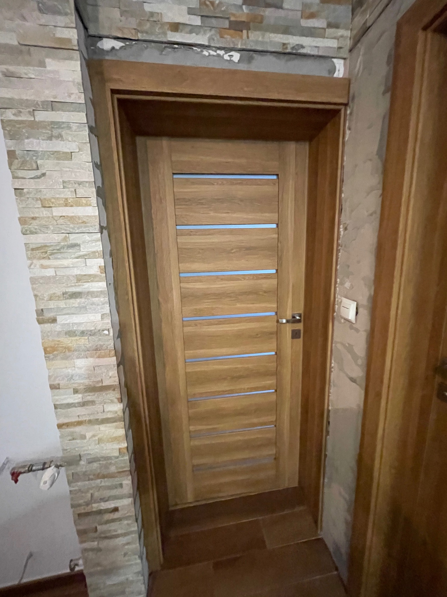 Nowoczesne, drewniane drzwi wewnętrzne z poziomymi przeszkleniami, osadzone w ościeżnicy. Obok dekoracyjna ściana z kamienia naturalnego i fragment ściany przygotowanej do malowania.