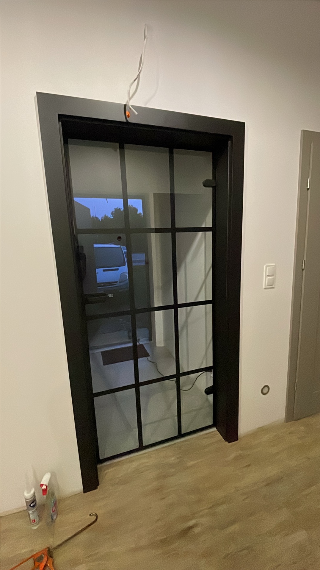 Czarne, przeszklone drzwi wewnętrzne z kratownicą, widoczne narzędzia i chemia budowlana na podłodze. W tle widoczny samochód dostawczy.