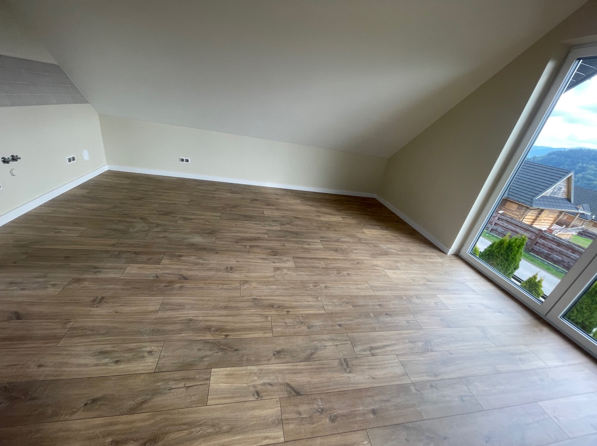 Jasne panele podłogowe w pokoju na poddaszu z oknem, przez które widać zieleń i góry. Ściany pomalowane na kremowo, widoczne gniazdka elektryczne.