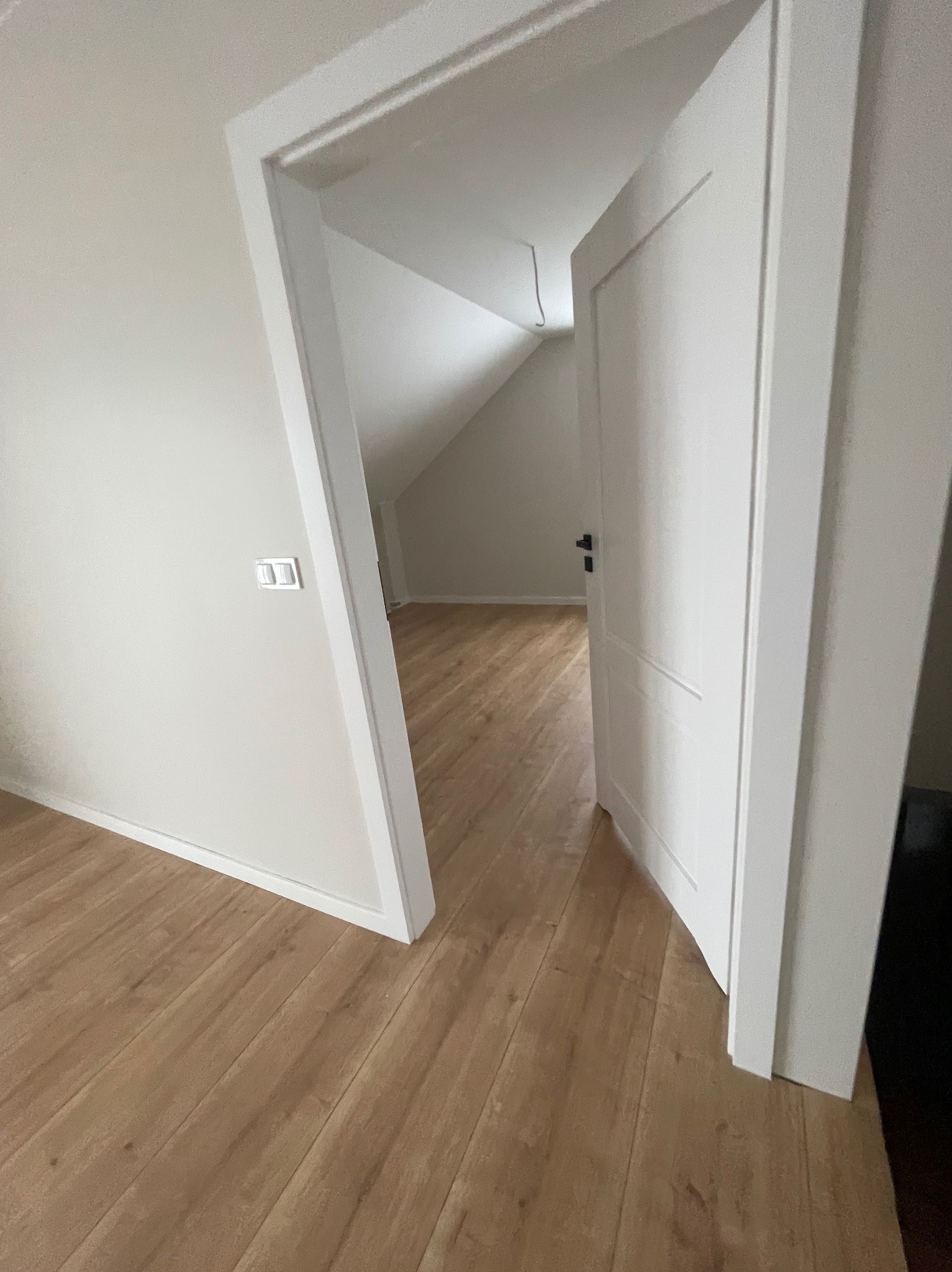 Jasne panele podłogowe w pokoju z otwartymi, białymi drzwiami prowadzącymi do poddasza. Ściany w odcieniach bieli, widoczny włącznik światła.