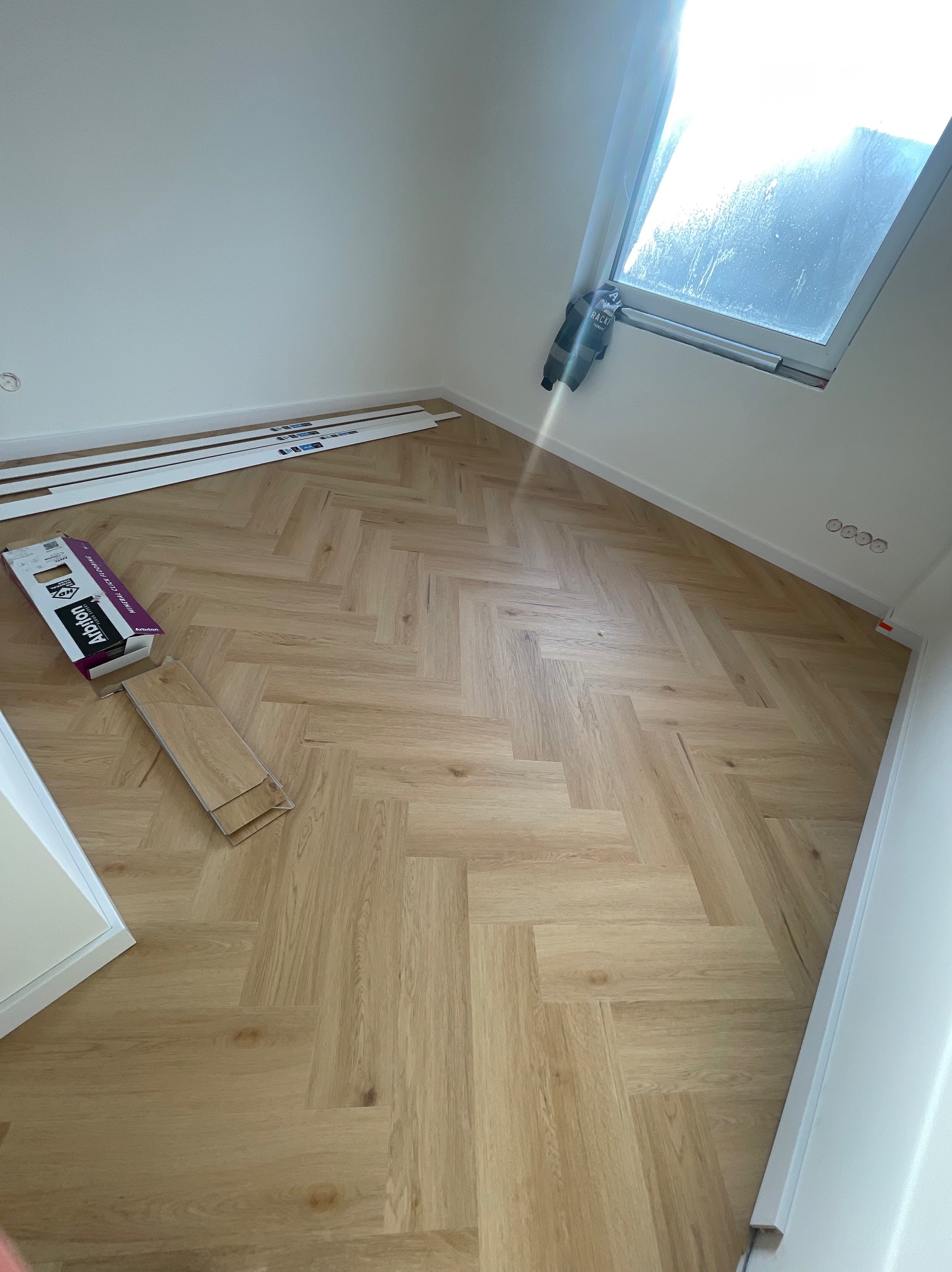 Ukończona podłoga z paneli ułożonych w jodełkę, widoczne listwy przypodłogowe i okno dachowe. Częściowo widoczne opakowanie paneli i kilka niezamontowanych desek.