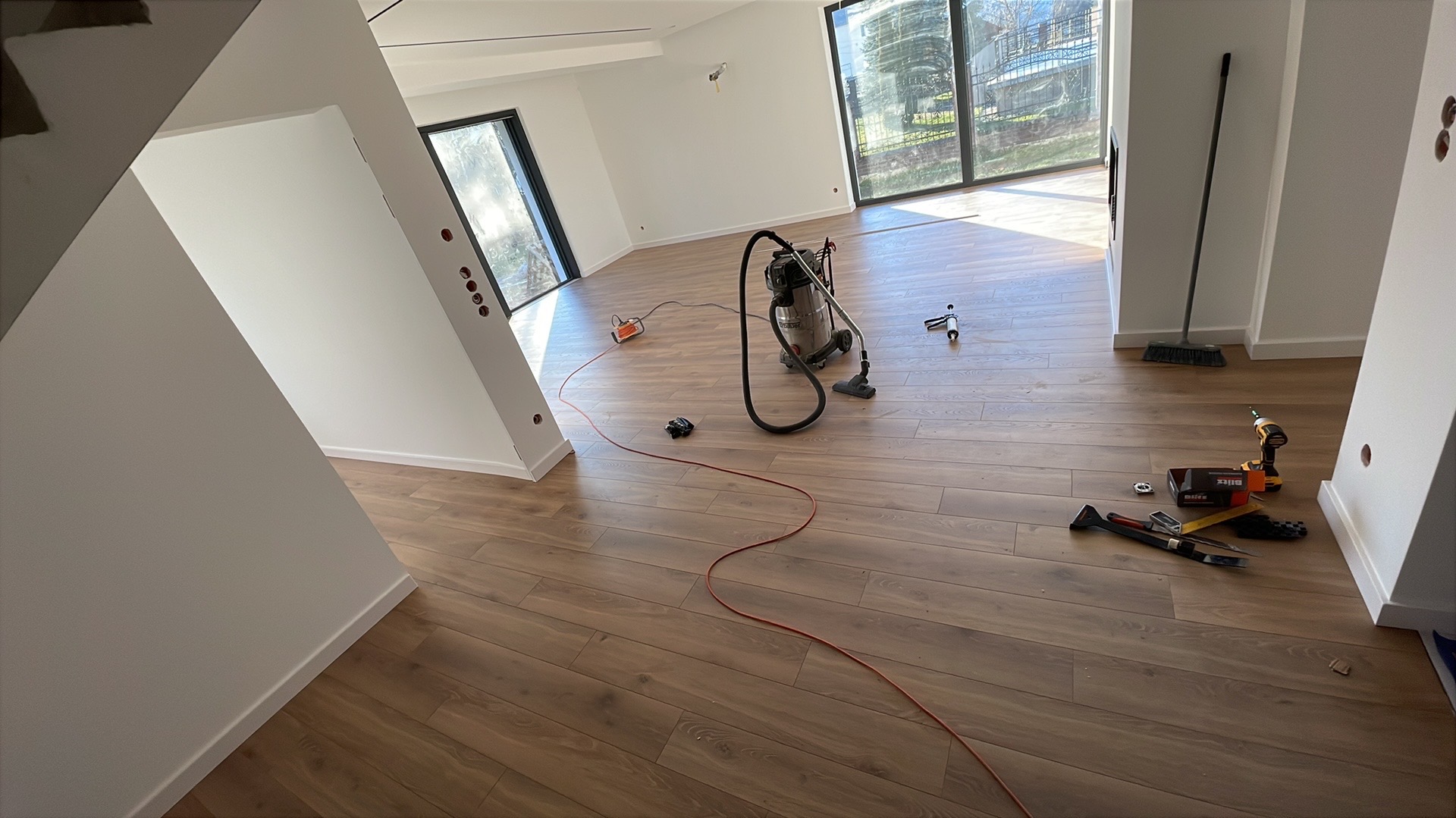Jasne wnętrze po ułożeniu paneli podłogowych. Widoczne narzędzia, odkurzacz i listwy przypodłogowe. Duże okna wpuszczają naturalne światło.
