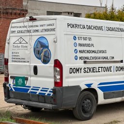 Voodoo Graphics - Tył białego samochodu dostawczego z reklamą firmy budującej domy szkieletowe, widoczne numery telefonów, adres strony internetowej oraz adresy e-mail, na dole leżą metalowe profile.