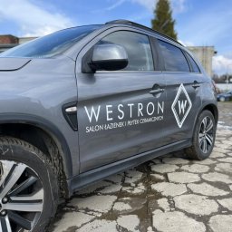 Voodoo Graphics - Szary SUV z logo i nazwą firmy 'Westron Salon Łazienek i Płytek Ceramicznych' na boku, zaparkowany na nierównym brukowanym podłożu.