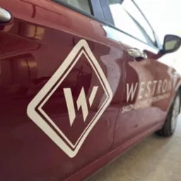 Bordowy samochód z białym logo firmy 'Westron Salon Lazienek' na drzwiach, ujęcie z boku, lekko z dołu.