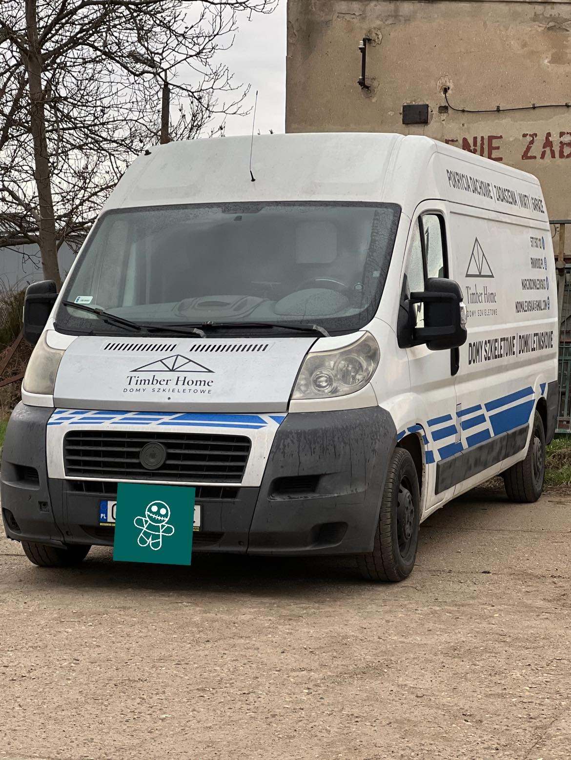 Biały samochód dostawczy firmy Timber Home z logo i niebieskimi pasami na karoserii, zaparkowany na szutrowej drodze przed budynkiem. Tablica rejestracyjna zasłonięta zielonym symbolem.