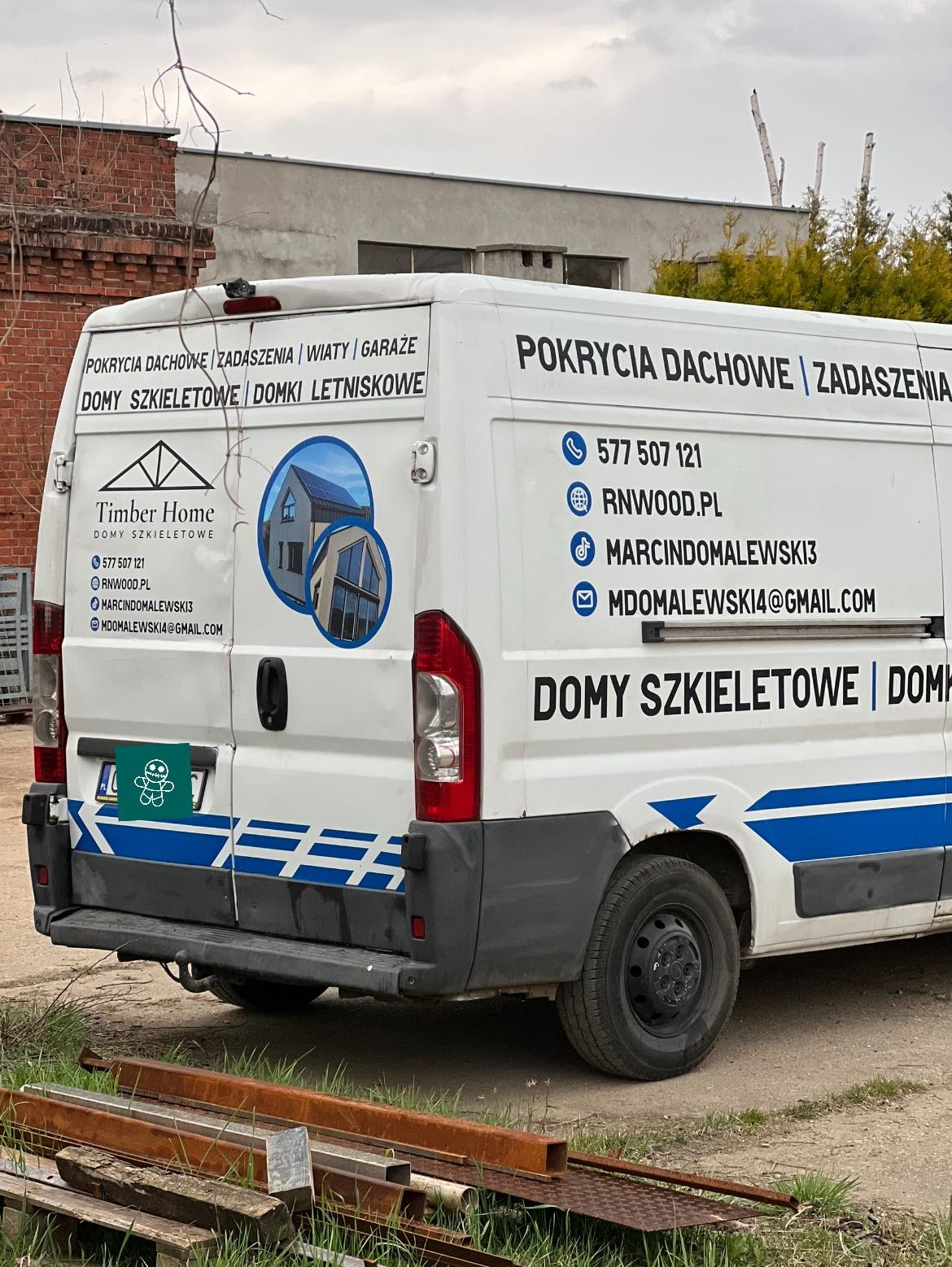 Tył białego samochodu dostawczego z reklamą firmy budującej domy szkieletowe, widoczne numery telefonów, adres strony internetowej oraz adresy e-mail, na dole leżą metalowe profile.