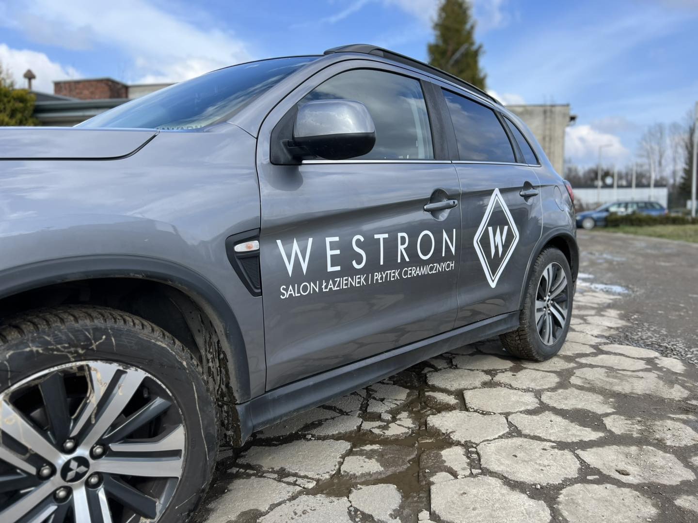 Szary SUV z logo i nazwą firmy 'Westron Salon Łazienek i Płytek Ceramicznych' na boku, zaparkowany na nierównym brukowanym podłożu.