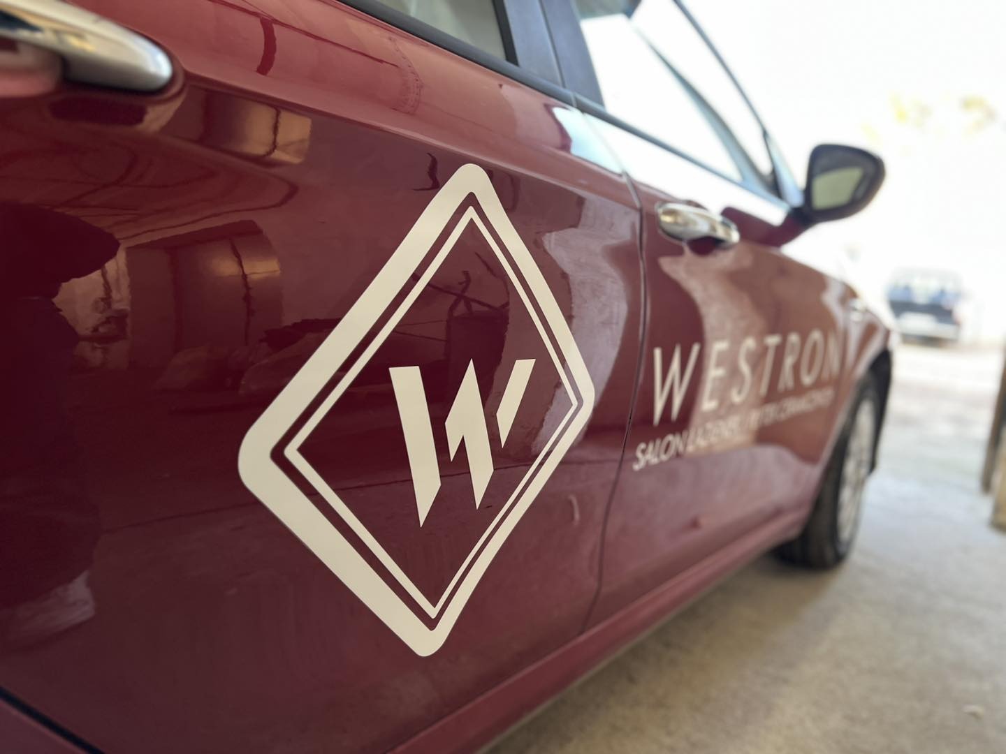 Bordowy samochód z białym logo firmy 'Westron Salon Lazienek' na drzwiach, ujęcie z boku, lekko z dołu.