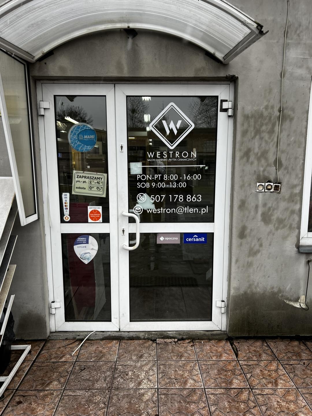 Wejście do salonu łazienek Westron z logo na szybie, naklejkami informacyjnymi i godzinami otwarcia, widoczne kafelki pod drzwiami.