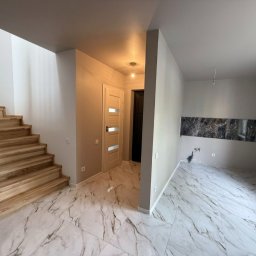 Andrzej Serwis - Jasne wnętrze domu z drewnianymi schodami i marmurową podłogą. Widoczne drzwi wewnętrzne z przeszkleniami i fragment ściany z dekoracyjnym pasem imitującym marmur.