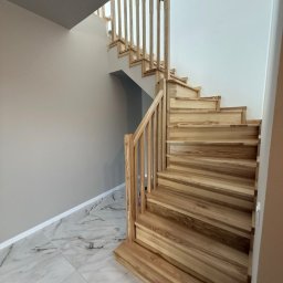 Andrzej Serwis - Jasne, drewniane schody z balustradą w nowoczesnym wnętrzu. Widoczne marmurowe płytki podłogowe i ściany w odcieniach szarości i bieli. Ujęcie z dołu, podkreślające konstrukcję.