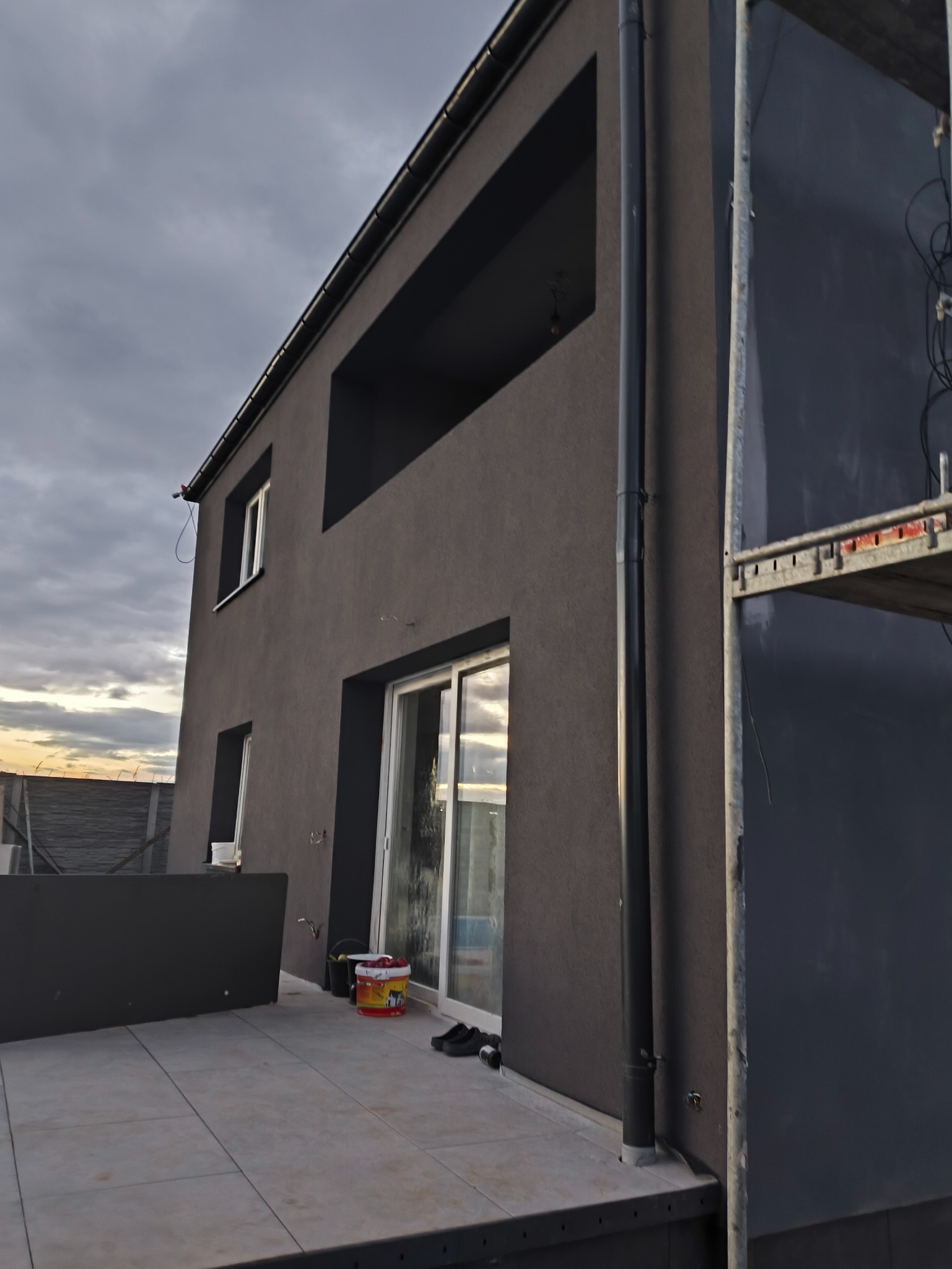 Elewacja domu w ciemnej tonacji z widocznymi rynnami i fragmentem rusztowania. Na pierwszym planie balkon z płytkami i akcesoriami malarskimi.