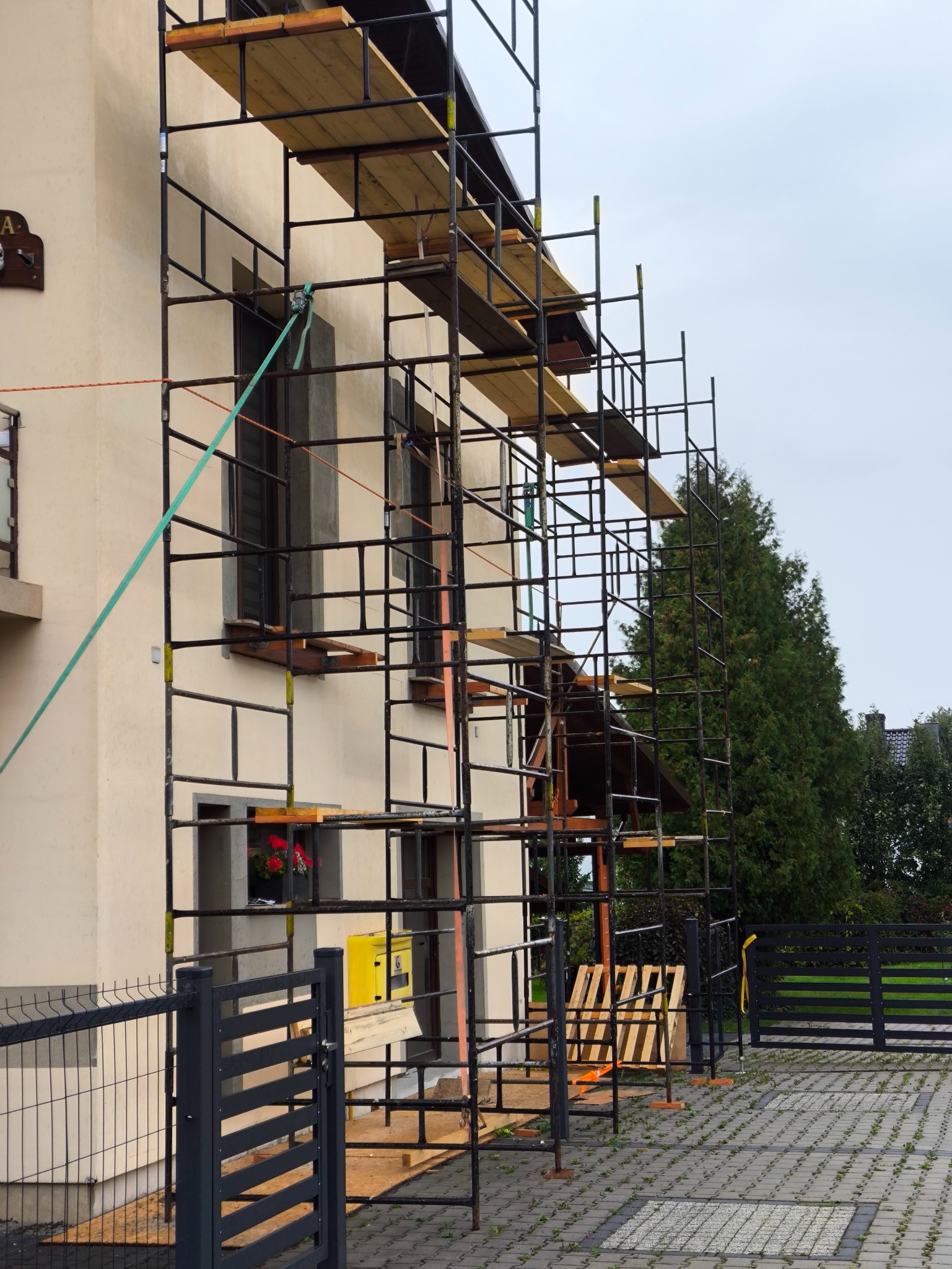 Dom w trakcie remontu, z rusztowaniem obejmującym elewację. Widoczne okna, fragment dachu i ogrodzenie. Prace budowlane w toku, z elementami wyposażenia na rusztowaniu.