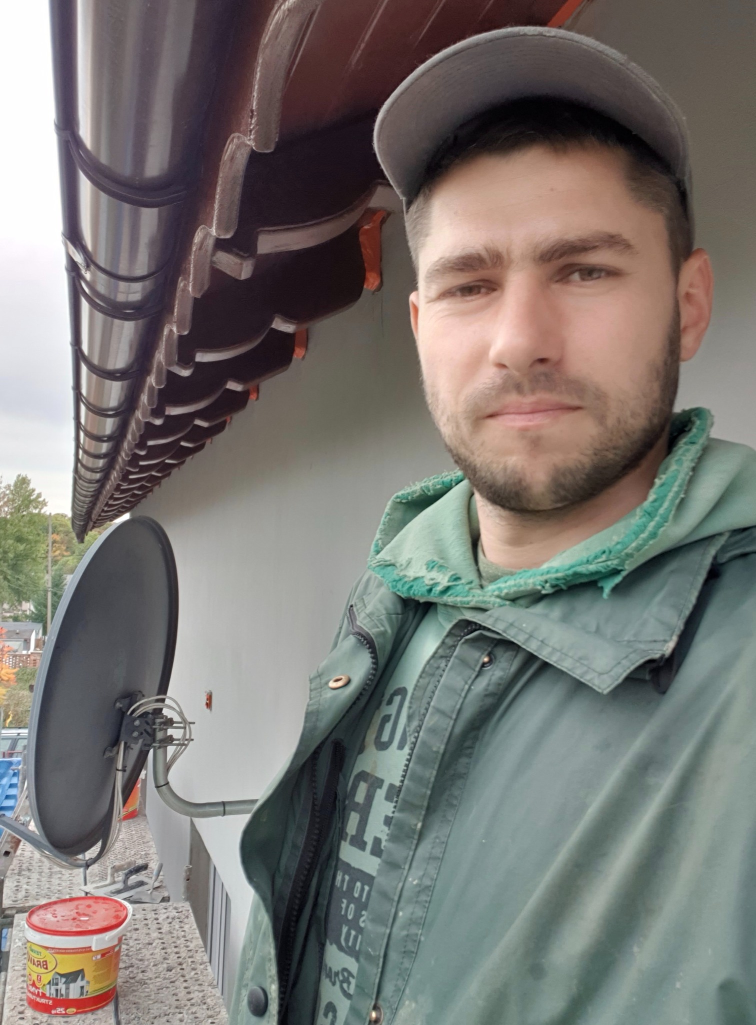 Mężczyzna w czapce i kurtce na rusztowaniu, obok brązowe rynny i antena satelitarna. Widoczny kubeł z farbą. Ujęcie z dołu.