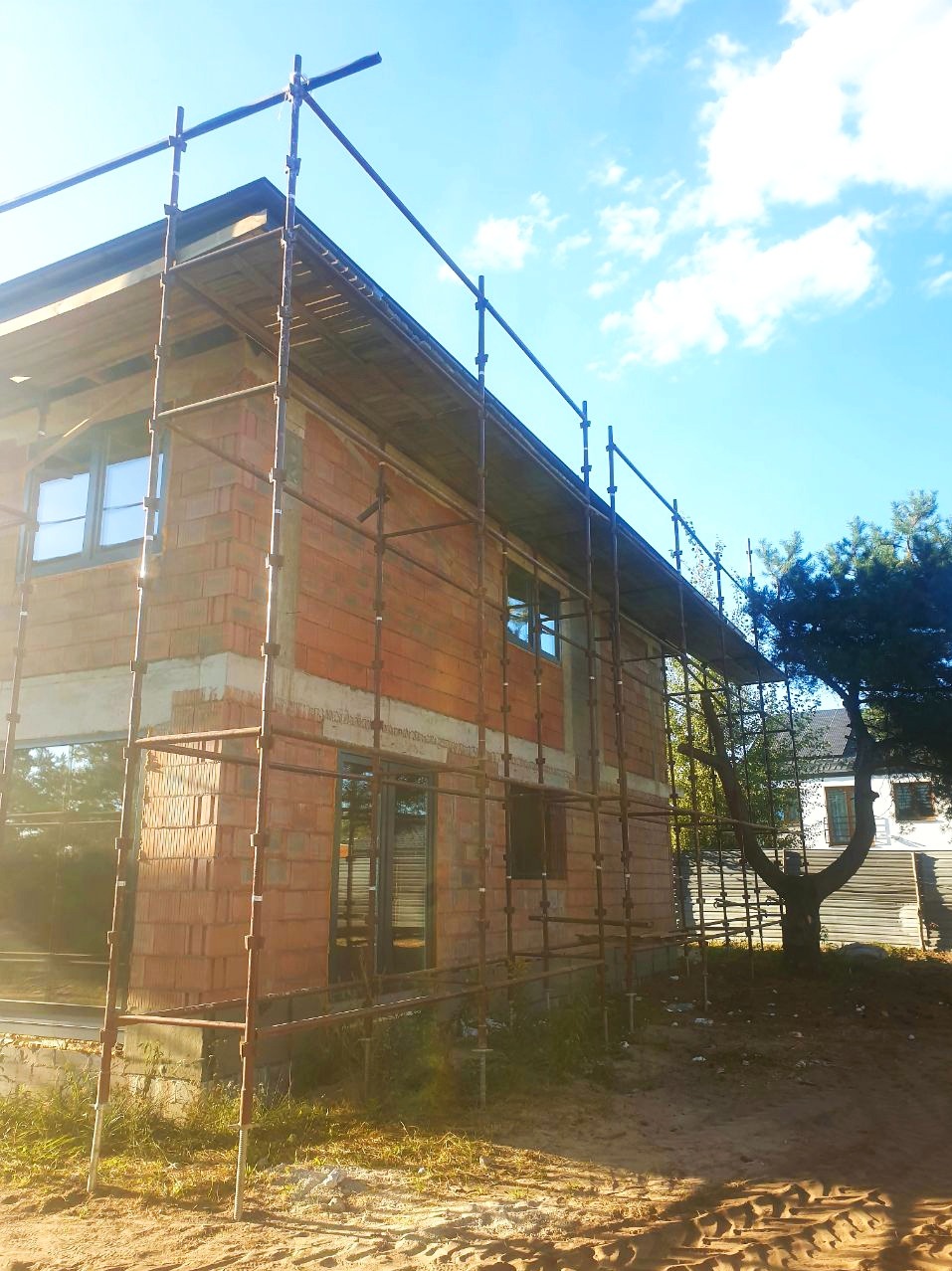 Niedokończony budynek z czerwonej cegły otoczony rusztowaniem pod błękitnym niebem. Widoczne okna i fragment drzewa. Ujęcie z dołu, podkreślające wysokość konstrukcji.