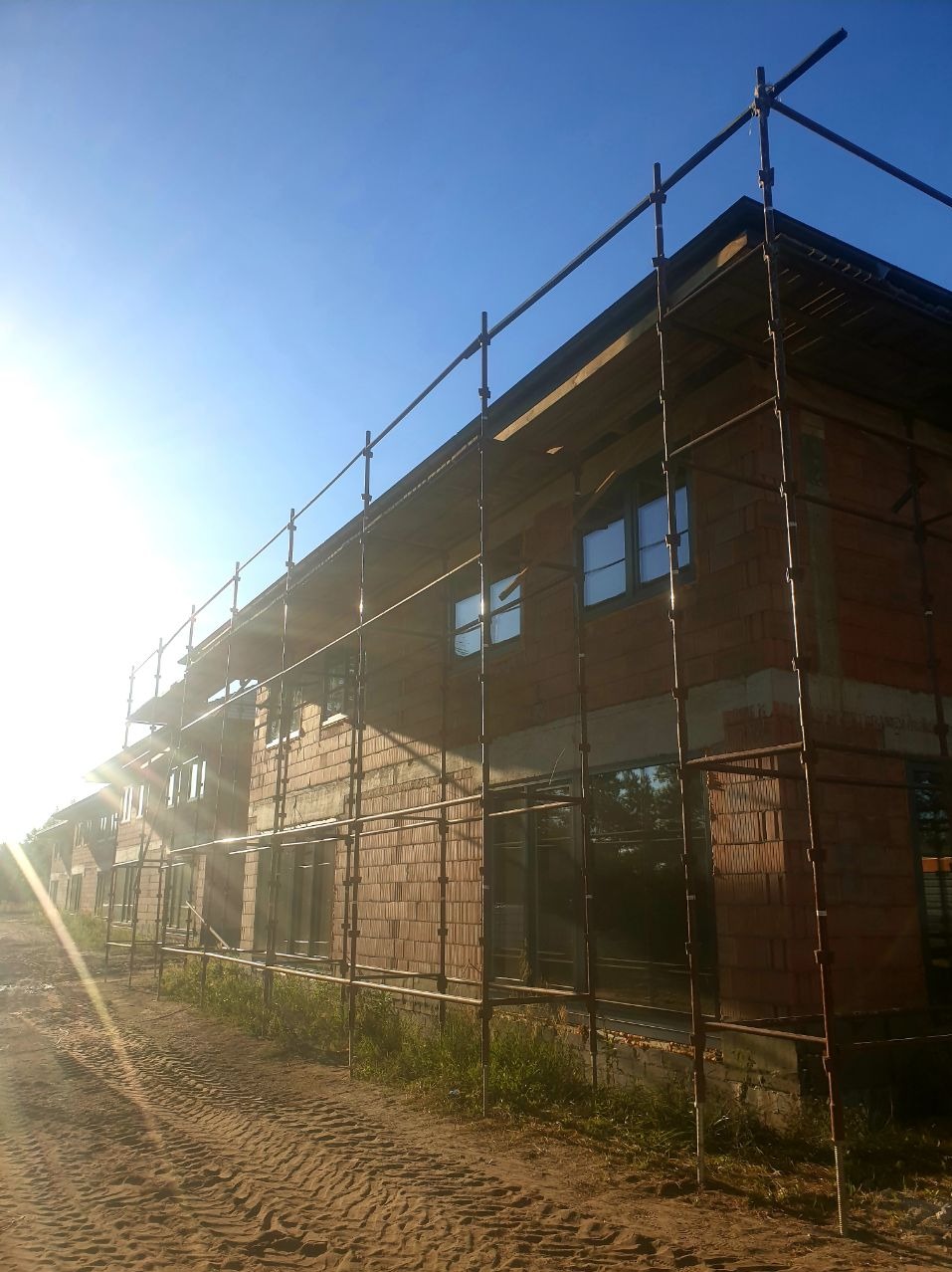 Budynek w trakcie budowy z cegły, otoczony rusztowaniem, w słoneczny dzień. Widoczne okna i fragment dachu. Prace murarskie w toku.