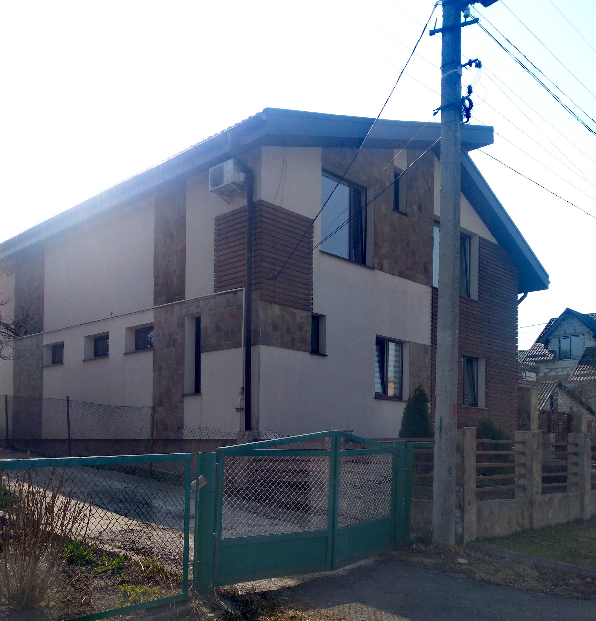 Dwukondygnacyjny dom z elewacją w odcieniach beżu i brązu, z elementami imitującymi kamień i drewno. Widoczny klimatyzator na ścianie i zielona brama wjazdowa.