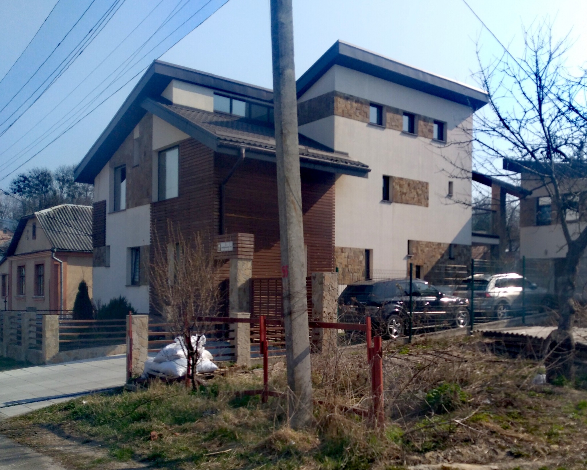 Dwupiętrowy dom z drewnianą elewacją i szarą dachówką, widoczny zza ogrodzenia. Na podjeździe zaparkowane dwa samochody. W tle drugi budynek.