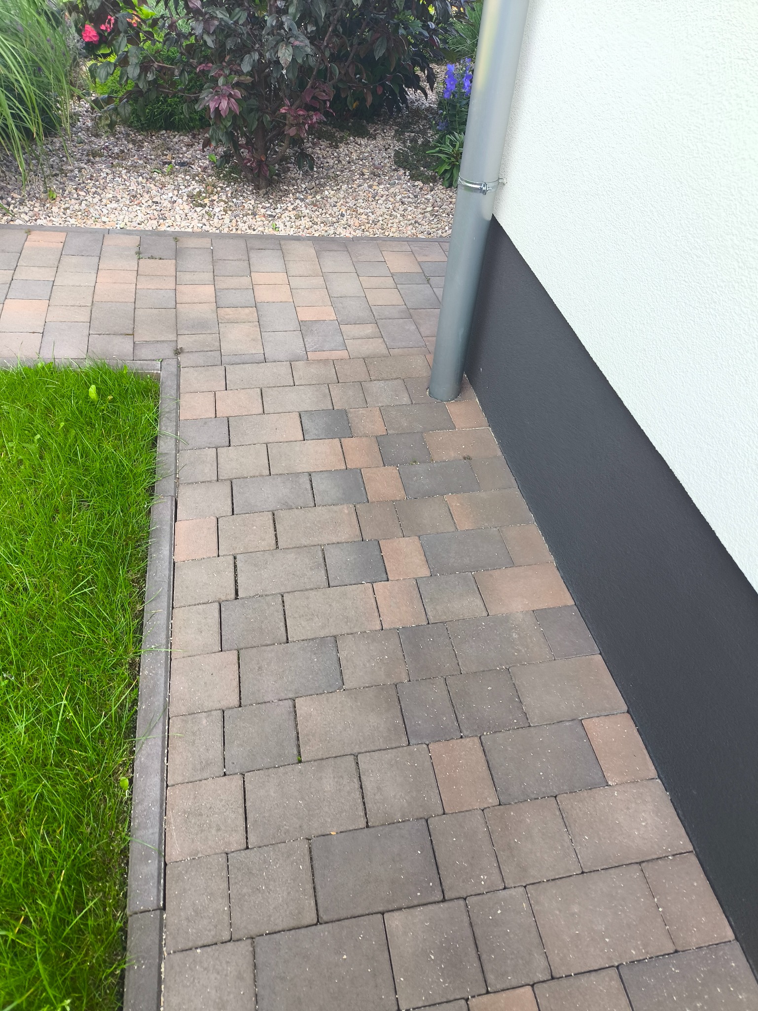Ułożona kostka brukowa w odcieniach brązu i szarości, przylegająca do ściany budynku z rurą spustową. Widoczny fragment trawnika i rabata z kamykami.