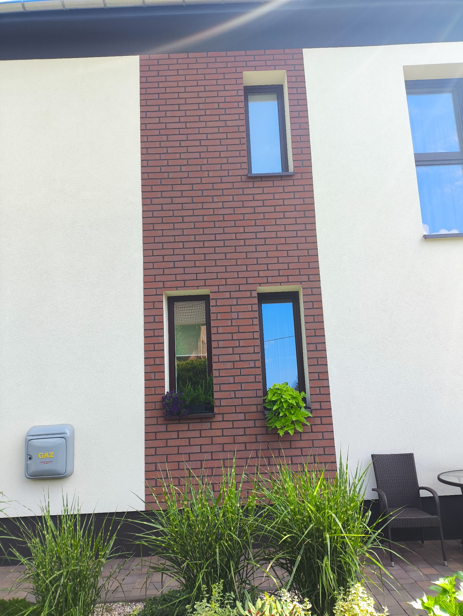 Elewacja domu z ceglaną wstawką, trzema oknami i skrzynkami z kwiatami. Widoczny licznik gazu i fragment tarasu z krzesłem.