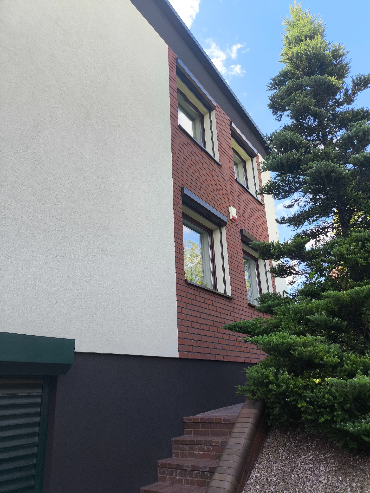 Elewacja budynku z ceglaną ścianą i białym tynkiem, widoczne okna z roletami, fragment schodów i iglaste drzewo. Ujęcie z dołu, błękitne niebo w tle.