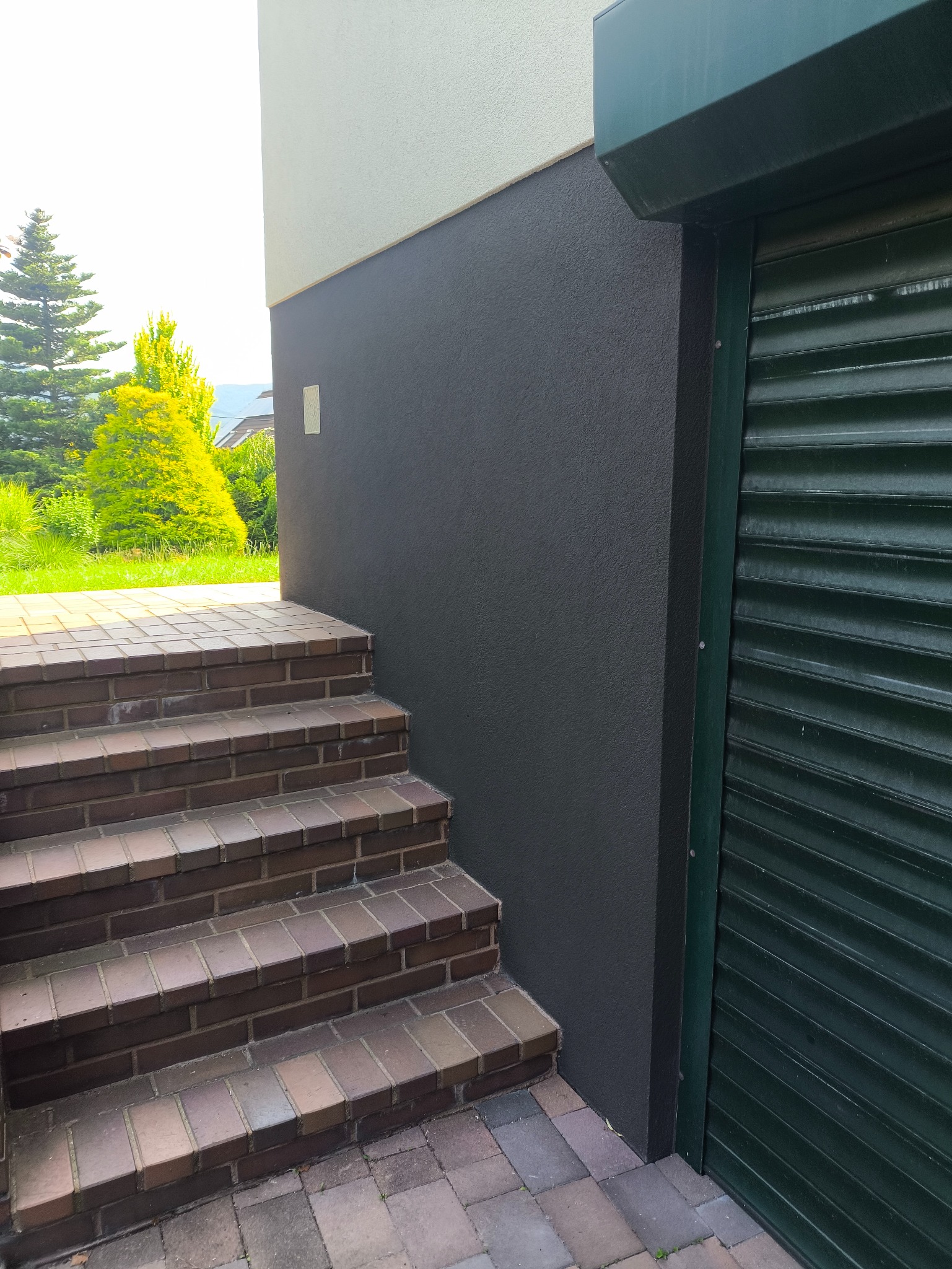 Schody z cegły klinkierowej prowadzące do budynku z szarą elewacją i zieloną roletą garażową. Widoczne drzewa w tle.