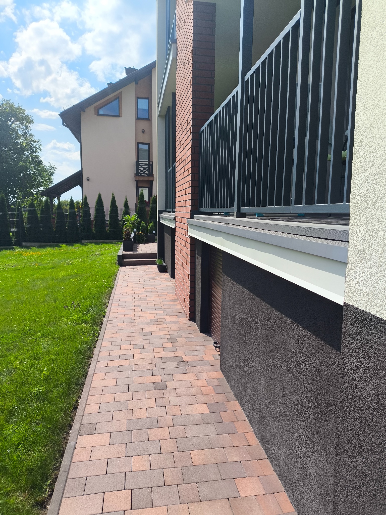 Nowoczesny dom z ceglaną elewacją i metalową balustradą balkonu, widok z poziomu chodnika z kostki brukowej. Zadbany trawnik i tuje w tle.