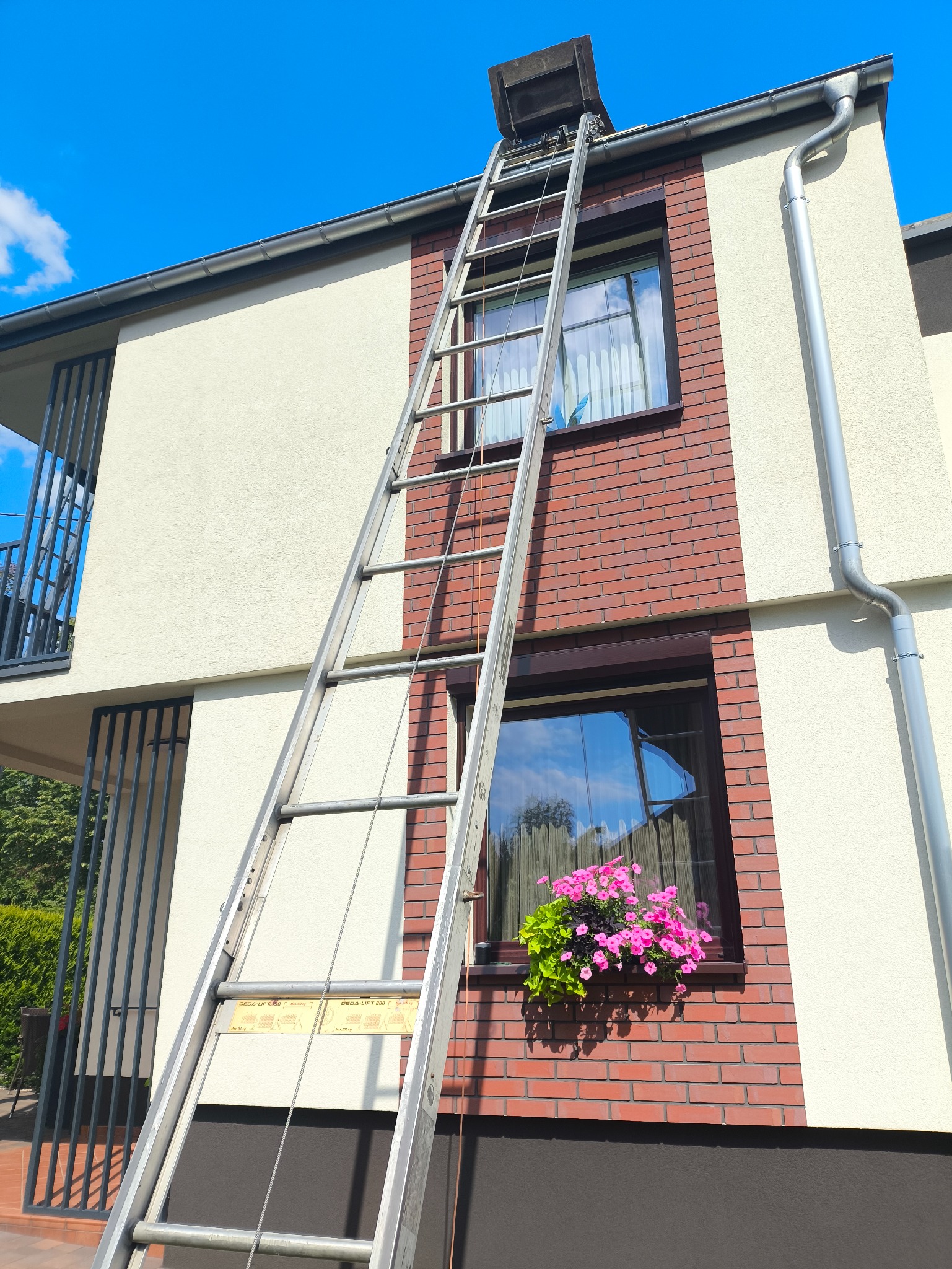 Aluminiowa drabina przystawiona do dwupiętrowego domu z ceglaną elewacją i rynnami. Na dachu widoczny właz. Okno ozdobione kwiatami w doniczce.