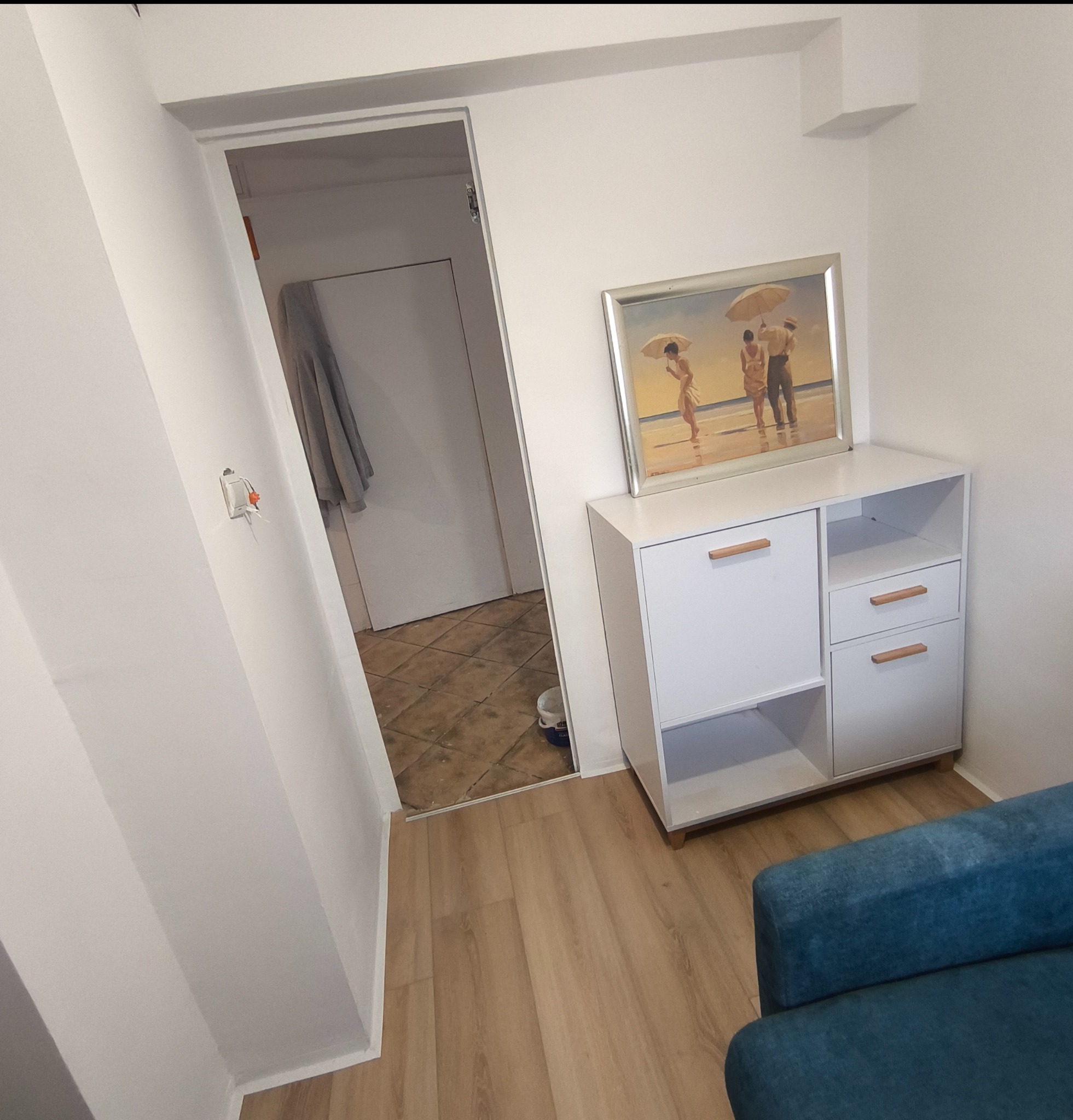Biała komoda z obrazem w pokoju po remoncie. Widoczny fragment niebieskiej sofy i otwarte drzwi do innego pomieszczenia z płytkami na podłodze.