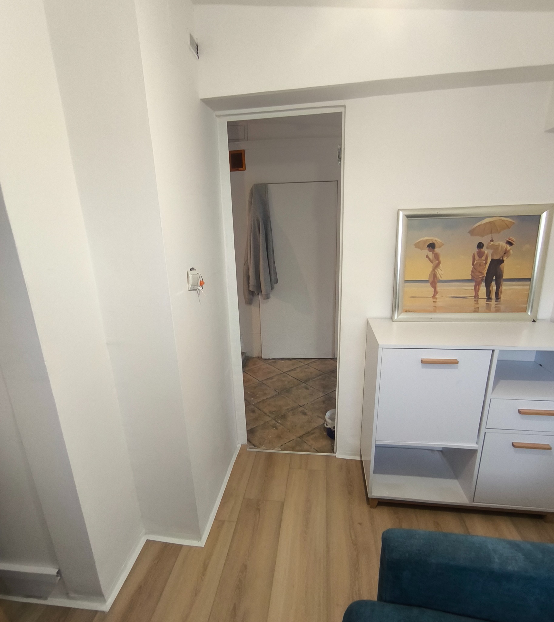 Białe ściany i podłoga w mieszkaniu. Widok na otwarte drzwi do innego pomieszczenia. Biała komoda z obrazem na górze. W tle widoczny fragment instalacji elektrycznej.