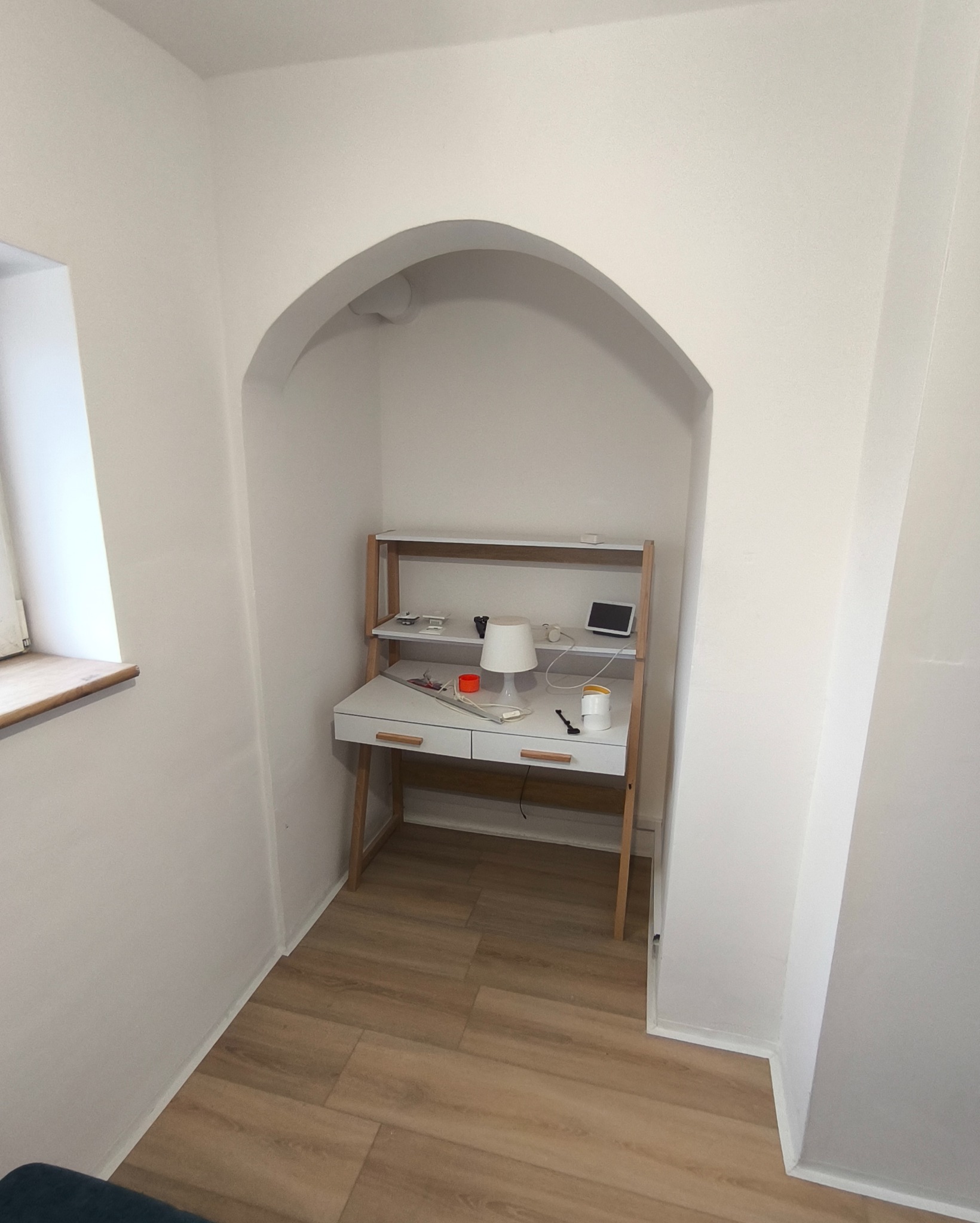 Białe biurko z drewnianymi nogami i półkami, umieszczone we wnęce z łukowym wejściem. Na blacie lampka, kubek i inne drobne przedmioty. Podłoga wyłożona panelami.